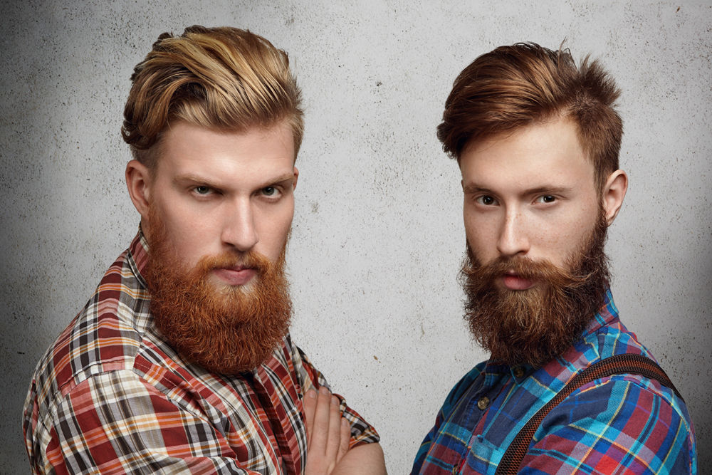 Two handsome young unshaved men with hipster beards dressed in stylish checkered shirts, standing with arms folded on gray concrete wall, looking with serious face expression, squinting eyes